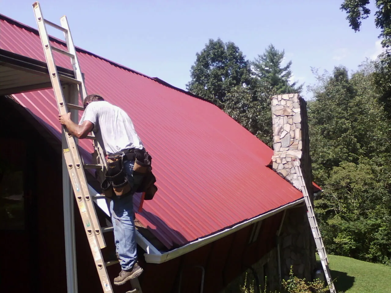 Roofer on a ladder beside a red metal roof and stone chimney — Soffit & Fascia Repair by Roofing Brothers in Avenue